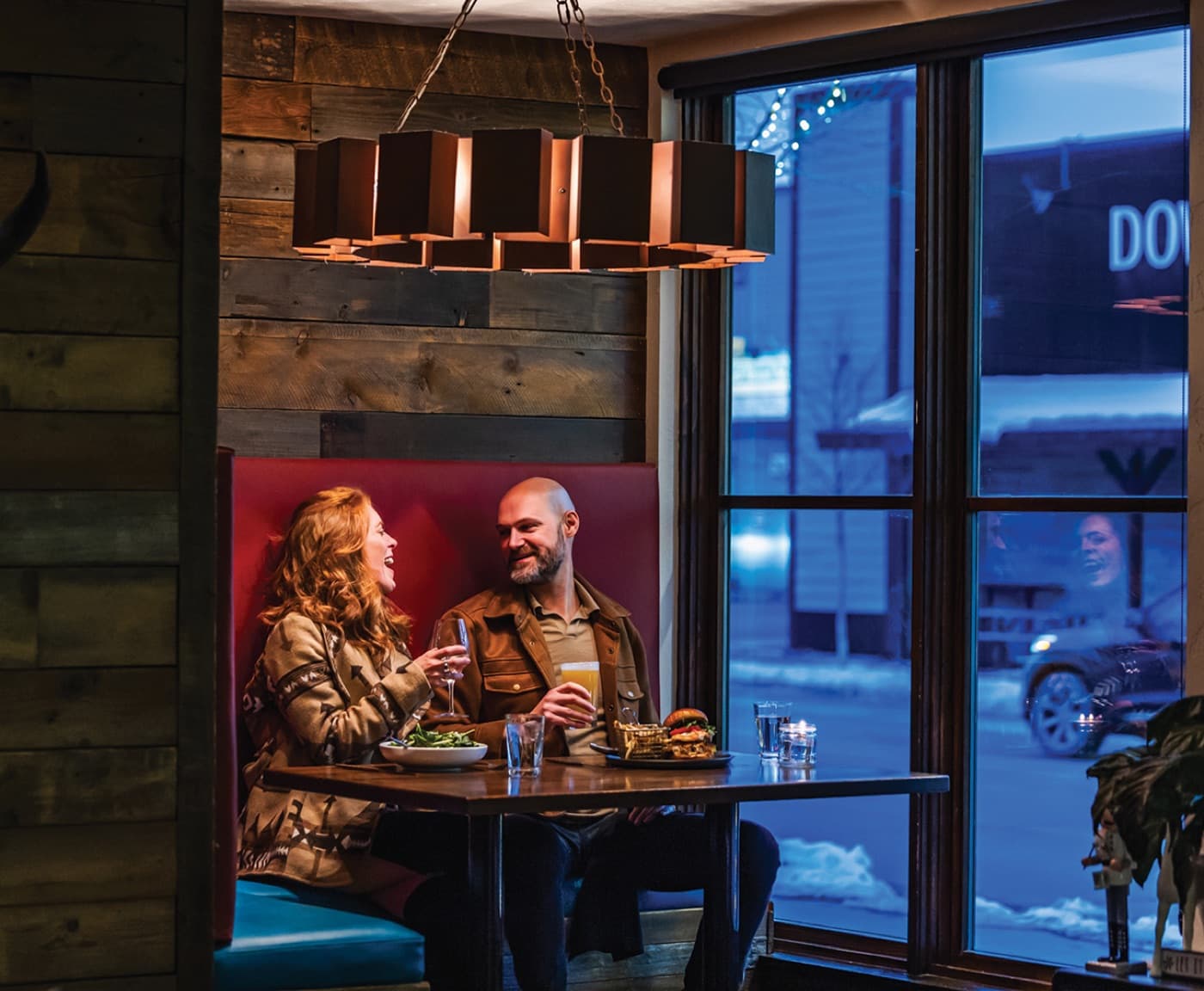 A couple laughing over dinner in a booth at The Firebrand's restaurant, snowy downtown Whitefish out the window