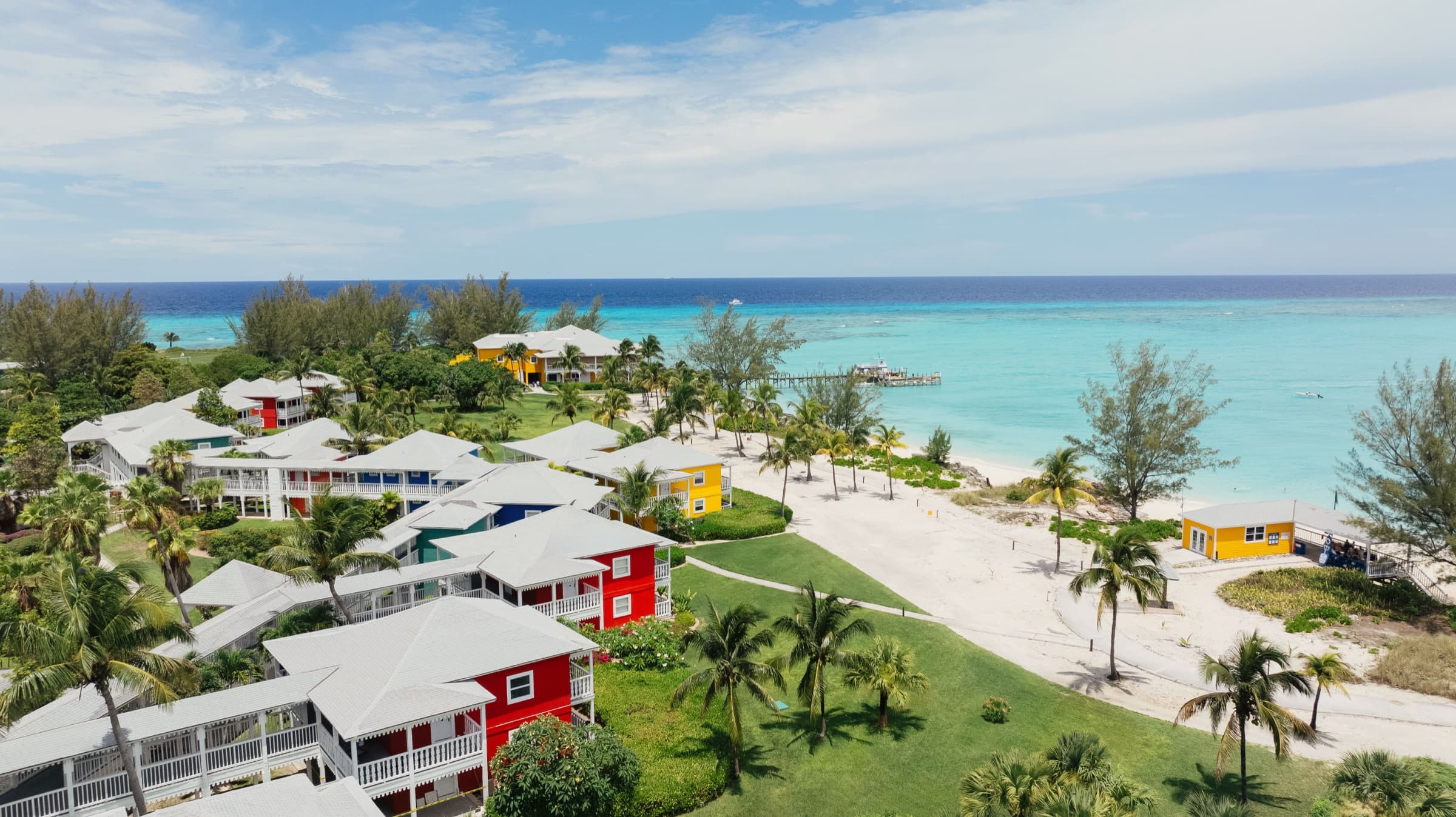 Aerial view of Club Med Columbus Isle — brightly colored bungalows among palms on a white-sand shore, turquoise water to the reef line