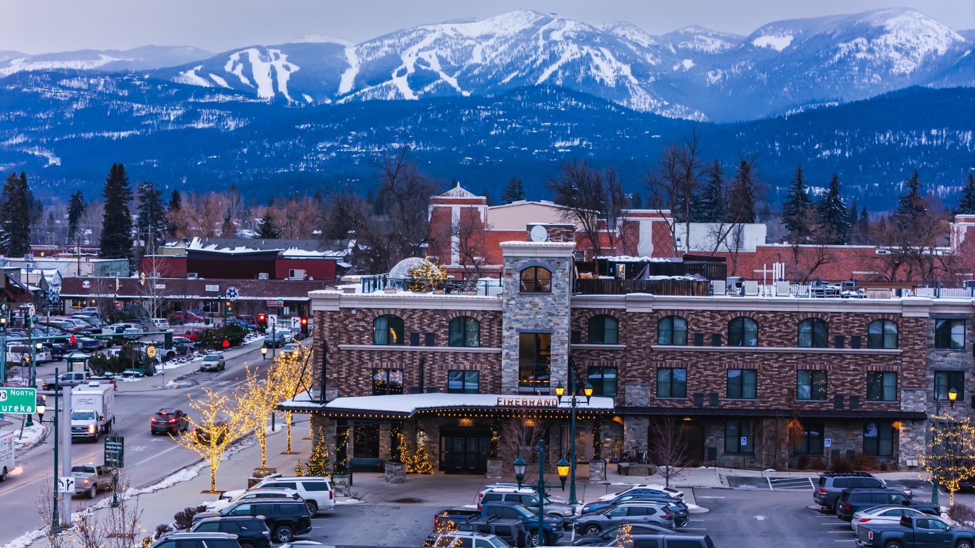 The Firebrand Hotel in downtown Whitefish at dusk, holiday lights glowing, the ski runs of Whitefish Mountain rising behind town