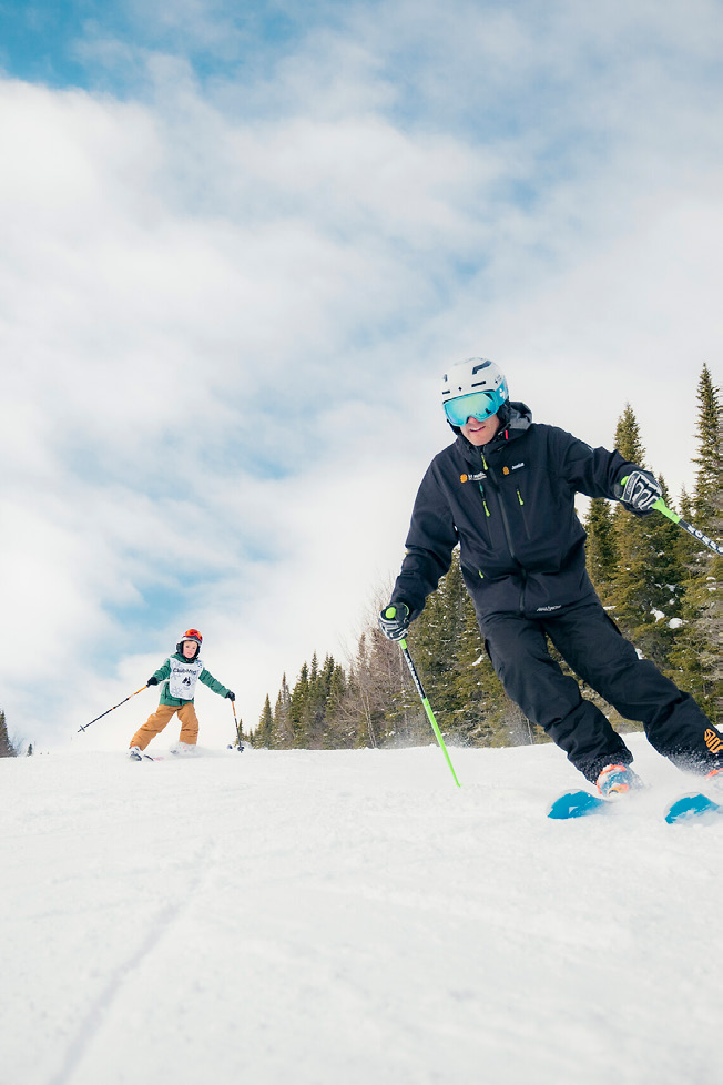 A Club Med ski instructor carving down a groomed Engadine run with a young skier following behind