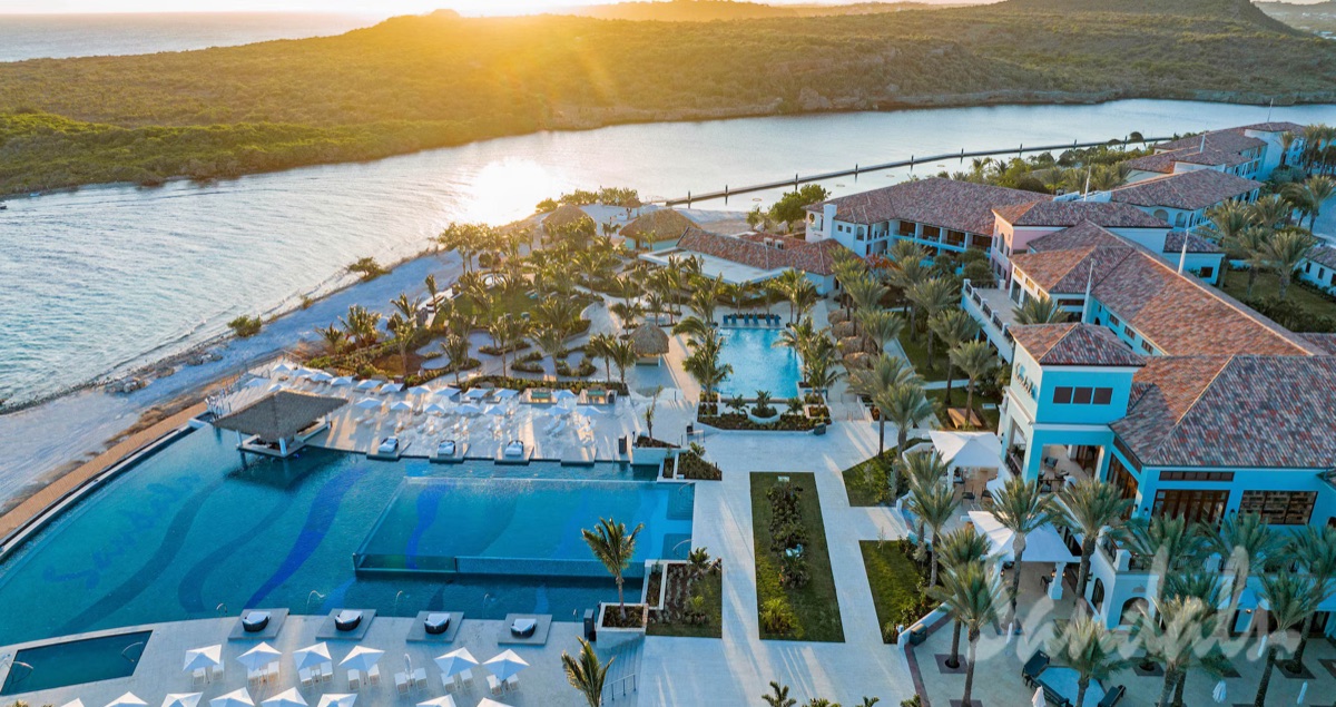 Sunset aerial over Sandals Royal Curaçao — the Dos Awa infinity pool, terracotta roofs, and the Spanish Water beyond
