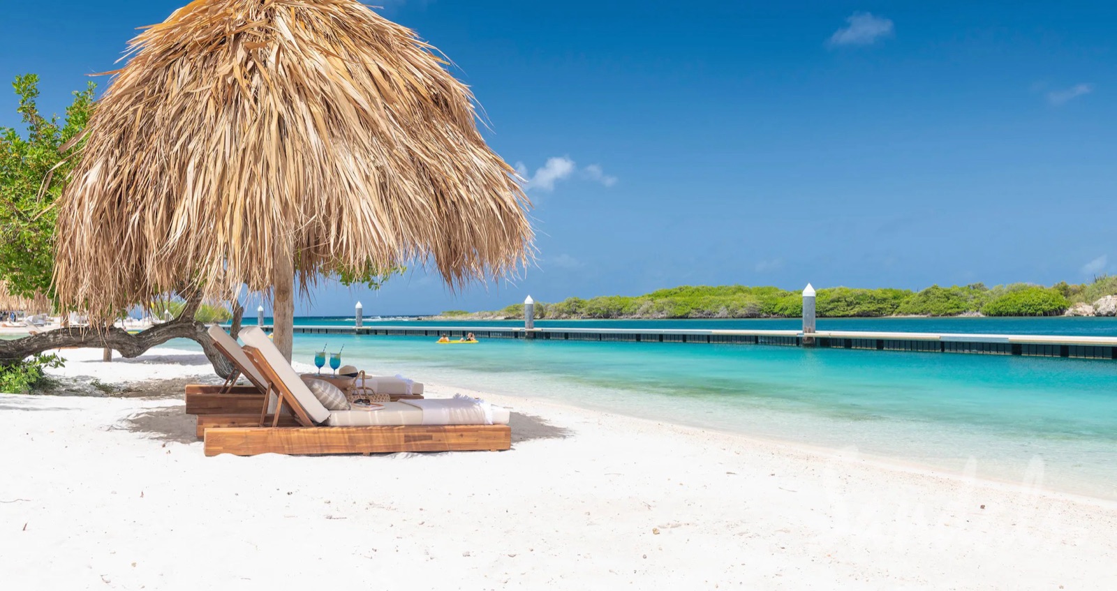 A thatched palapa and loungers on the white-sand beach at Sandals Royal Curaçao, with the floating pier beyond