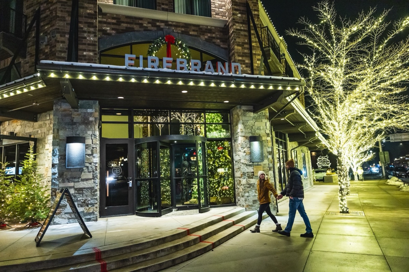 The Firebrand Hotel entrance at night, snow on the awning and the sidewalk trees wrapped in lights