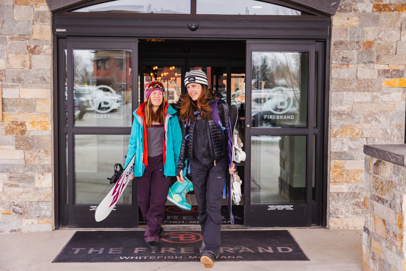 Two snowboarders walking out of The Firebrand Hotel's front doors, boards under their arms